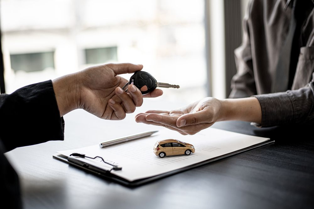 Two individuals discussing a car sale over a desk with one handing the other a set of car keys.